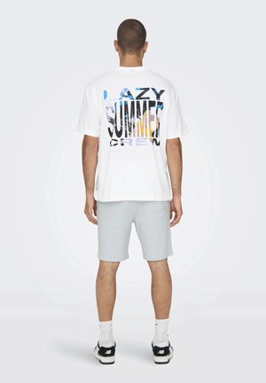 Mies, joka pitää valkoista T-paitaa, jossa on teksti "LAZY SUMMER CREW" ja grafiikkaa takaosassa, vaaleanharmaita shortsia, valkoisia sukkia ja mustia New Balance -kenkiä.