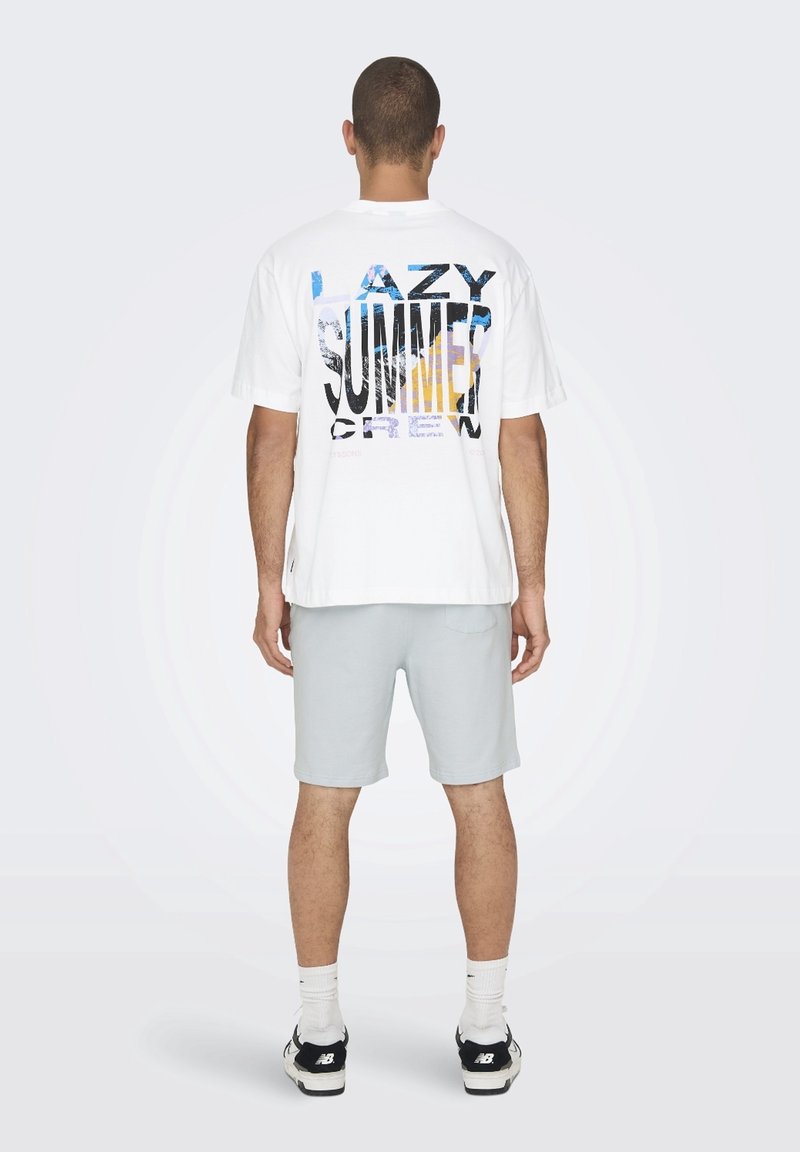 Mand iført en hvid T-shirt med teksten "LAZY SUMMER CREW" og grafik på ryggen, lysegrå shorts, hvide sokker og sorte New Balance-sko.