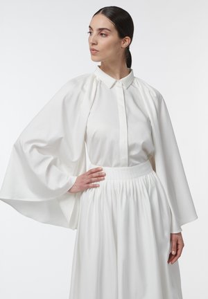 Femme aux longs cheveux noirs attachés en queue de cheval lisse, portant une tenue blanche fluide à manches larges et posant une main sur la hanche.
