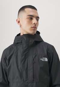 Svart vattentät jacka med hög krage, frampocket med dragkedja och broderad logotyp. Slät textur med minimal detaljering.