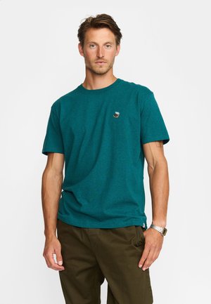 Camiseta de algodón color teal, con cuello redondo y mangas cortas, presenta un pequeño logo bordado en el pecho izquierdo. Combinada con pantalones verde oliva.