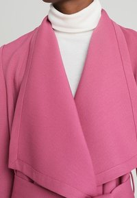 Rosa Wickelmantel mit weicher Textur, mit einem drapierten Vorderteil und Kragen, kombiniert mit einem weißen gerippten Rollkragenpullover.
