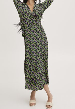 Femme portant une longue robe portefeuille noire à motif floral vert et violet, debout les bras levés, avec des sandales ouvertes beige à enfiler.