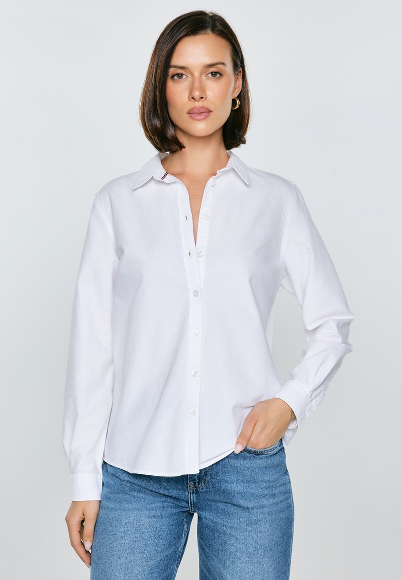 Femme aux cheveux courts bruns portant une chemise blanche à boutons et un jean bleu, debout avec une main dans la poche sur un fond clair.