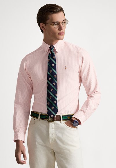 Camicia a righe rosa chiaro con colletto con bottoni, abbinata a una cravatta blu navy con motivo di stemmi. Pantaloni beige e cintura multicolore completano il look.
