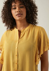 Gelbe Bluse mit Knopfleiste und kurzen, gerafften Ärmeln. Der Stoff hat einen dezenten Glanz und enthält vertikale Streifen für Textur und visuelles Interesse.