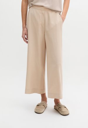 Person trägt beige, weitgeschnittene, gekürzte Hose und passende beige Slipper, steht mit einer Hand in der Tasche.