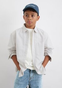 Camicia a righe grigia chiaro, indossata sopra una t-shirt bianca, abbinata a jeans di denim blu chiaro, cappellino da baseball blu scuro con visiera piatta.