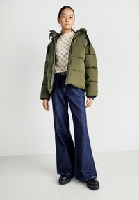 Giacca imbottita verde oliva con cappuccio, abbinata a una maglietta con motivo di pizzo e jeans a gamba larga in denim blu scuro. Stivali platform neri chunky.