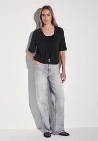 Cardigan nero a maglia con maniche corte sopra una maglietta nera, abbinato a jeans larghi grigio chiaro e ballerine in rete nere.
