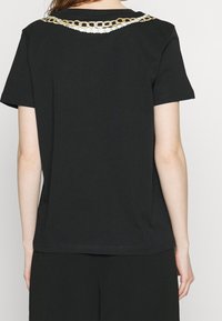 Svart t-shirt med rundad halsringning, med ett dekorativt kedjemönster i guld och vitt över axlarna. Korta ärmar, mjukt tyg.