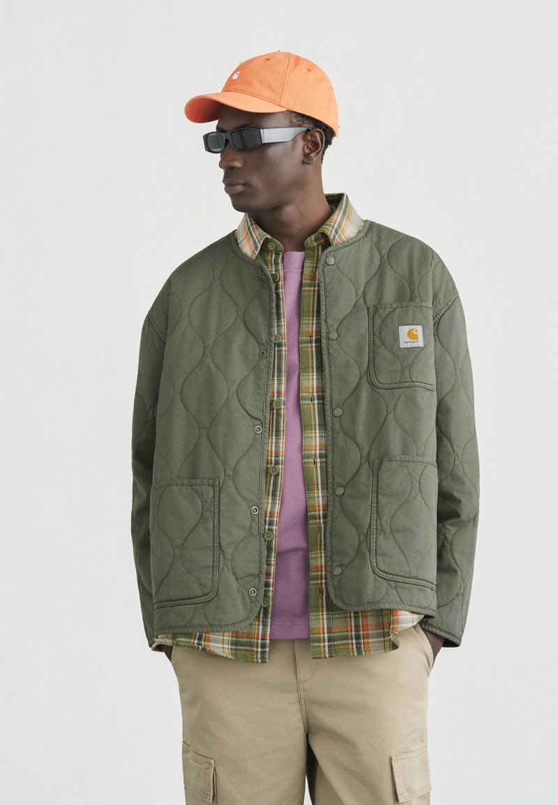 Homme portant une casquette orange, des lunettes de soleil noires, une veste matelassée verte sur une chemise à carreaux verte et un t-shirt violet, avec un pantalon cargo beige, regardant vers la gauche.