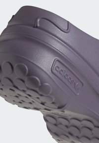 Zapatilla de goma morada con una superficie texturizada, patrón de suela redondeado y un logo grabado en el lateral.