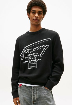 Pullover nero lavorato a maglia con testo del logo bianco e nomi di città. Collo e polsini a coste, abbinato a jeans grigi.