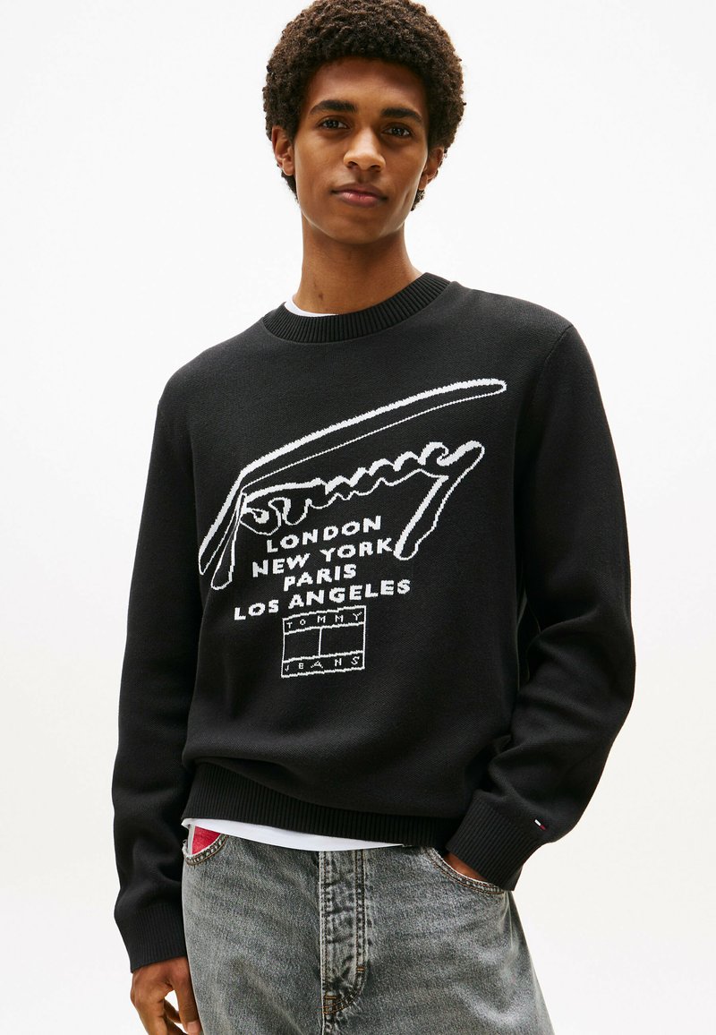 Schwarzer Strickpullover mit weißem Logo-Text und Städtenamen. Rippkragen und -manschetten, kombiniert mit grauen Jeans.