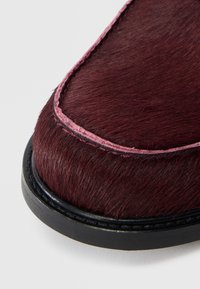 Close-up van een bordeauxrode schoentip met een textuur die lijkt op bont, roze stiksels en een zwarte rubberen zool op een witte achtergrond.