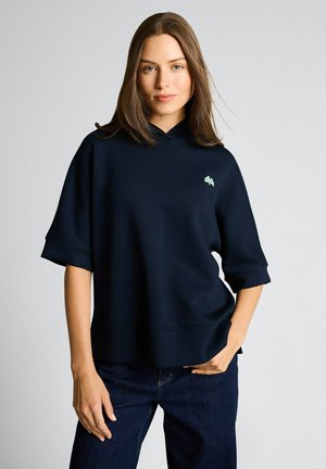 Kobieta o prostych brązowych włosach nosi luźną ciemnogranatową bluzę z krótkim rękawem i ciemne dżinsy, stoi na tle jednolitego, jasnego tła.