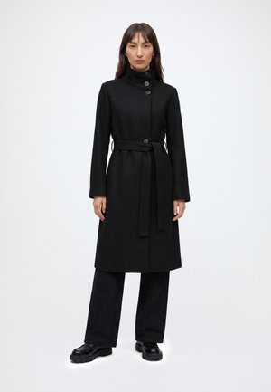 Femme portant un long manteau noir avec un col montant, boutonné devant, ceinture en tissu, pantalon noir et chaussures noires épaisses, debout sur un fond blanc.