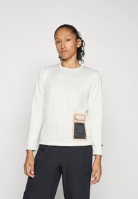 Top de manga larga color crema con tejido texturizado, que presenta un bolsillo marrón con un panel de malla en el lateral; combinado con pantalones oscuros.