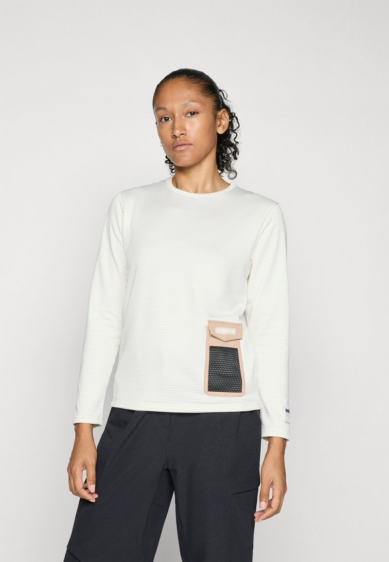 Top de manga larga color crema con tejido texturizado, que presenta un bolsillo marrón con un panel de malla en el lateral; combinado con pantalones oscuros.