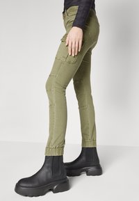Oliivivihreät cargo-housut sivutaskuilla, slim fit -mallia, ribatut nilkkapitsit, yhdistettynä mustiin tukeviin nilkkasaappaisiin.