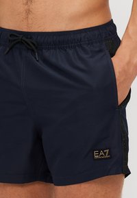 Shorts de bain bleu marine avec taille élastique, panneaux latéraux contrastés et accent de logo doré. Fabriqués en matériau léger et à séchage rapide.