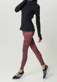 Svart långärmad träningsöverdel med hög krage, kombinerad med röda mönstrade leggings och svarta löparskor med vita detaljer.