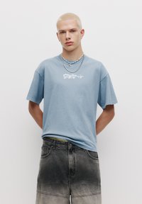 Tricou din bumbac de culoare albastru deschis, cu "STW" imprimat în alb, asociat cu pantaloni scurți din denim gri, cu efect ombre și detalii vizibile de cusătură.
