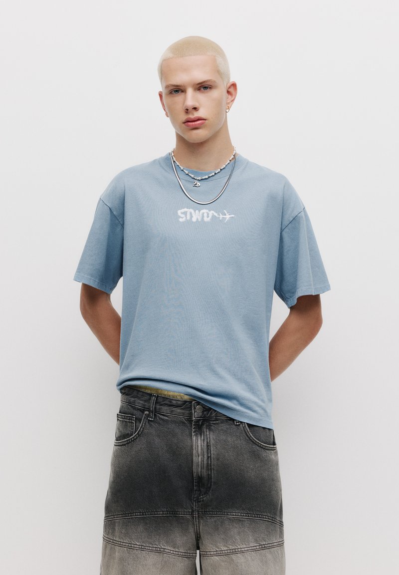 Tricou din bumbac de culoare albastru deschis, cu "STW" imprimat în alb, asociat cu pantaloni scurți din denim gri, cu efect ombre și detalii vizibile de cusătură.