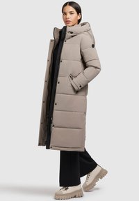 Lys grå lang pufferjakke med hætte, lynlås-lukning og sidelommer. Textureret stof med vandret quiltet mønster. Kombineret med chunky beige sko.