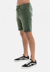 Shorts en denim vert avec des retroussages, design à cinq poches et une fermeture à bouton, associés à des baskets noires avec des accents blancs.