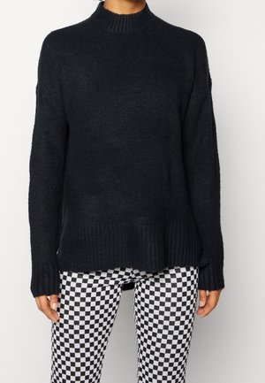 Person iført en sort langærmet sweater og sort-hvide ternede bukser, stående mod en ensfarvet baggrund.
