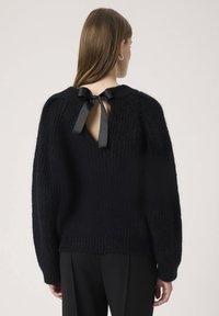 Pulover negru tricotat, cu o croială largă, textură ribbed și decolteu rotund. Prezintă un detaliu cu fundiță la spate și mâneci lungi.