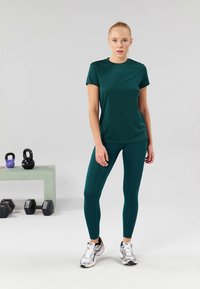Zelena sportska majica i izjednačene tajice od glatkog, rastezljivog materijala. Model nosi srebrne sportske cipele. U pozadini se nalaze tegovi i kettlebelli.