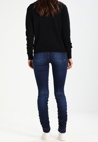 Svart texturerad tröja med ribbade ärmar, matchad med tajta mörkblå jeans och vita sneakers, sedd bakifrån.