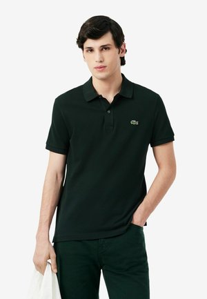 Hombre joven con una camiseta polo verde oscuro con un pequeño logo de cocodrilo, sosteniendo un paño blanco en una mano y con la otra mano en el bolsillo.