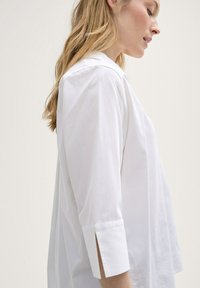 Chemise blanche à manches longues avec une coupe décontractée, manches 3/4 et fentes latérales au niveau des poignets. Tissu en coton doux, design minimaliste.