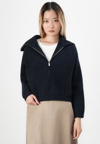 Jeune femme aux cheveux blonds raides portant un sweat à capuche navy par-dessus une chemise blanche et une jupe beige, se tenant contre un fond blanc.