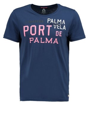 Mand går med navyblå grafisk T-shirt, lyse beige cargoshorts og grå sneakers på ensfarvet lys baggrund.