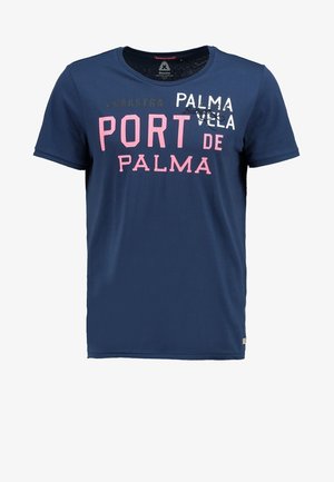 Mężczyzna idzie, nosząc granatowy T-shirt z nadrukiem, jasnobeżowe krótkie spodnie typu cargo i szare sneakersy na jednolitym jasnym tle.