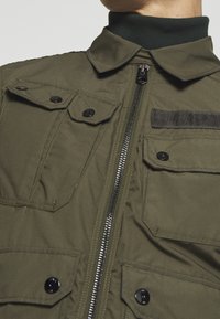 Olijfgroene jacket met een rits aan de voorkant, twee klepzakken en een zwarte accentstrip. Beschikt over een kraag en een geribbelde groene nekzone.