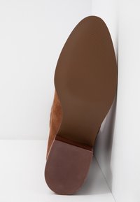 Semelle d'une chaussure en daim marron avec talon en caoutchouc texturé et avant-pied en caoutchouc lisse, photographiée sur fond blanc.