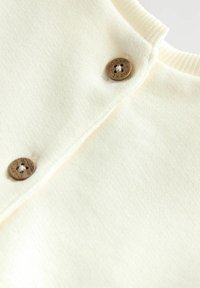 Krämfärgad stickad tröja med detaljer som en ribbad krage och två mörkbruna knappar med fyra hål och graverad text.