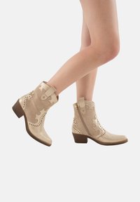 Botines beige que combinan texturas lisas y de ante, con recortes decorativos, detalles con clavos y un tacón bajo y cuadrado.