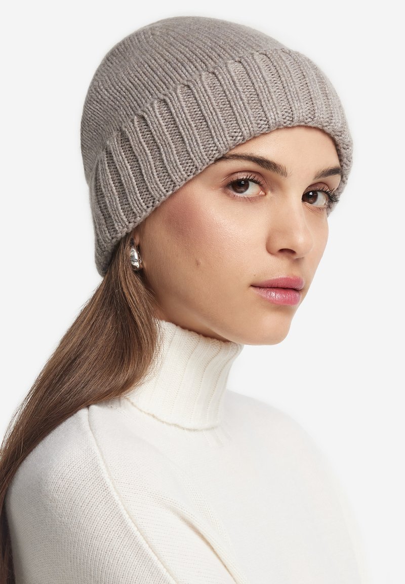 Frau mit langen braunen Haaren, die eine graue Strickmütze, silberne Creolen und einen weißen, gerippten Rollkragenpullover trägt und seitlich schaut.