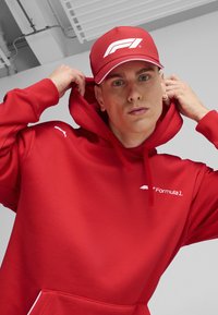 Hanorac roșu cu buzunar frontal și glugă ajustabilă; prezintă logo-ul alb "Puma" și textul "Formula 1". șapca roșie afișează logo-ul alb F1.