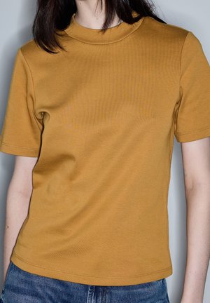 Osoba z ciemnymi włosami ubrana w zwykły musztardowożółty krótki rękaw t-shirt i niebieskie dżinsy na neutralnym tle.