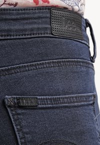 Vaqueros de mezclilla azul oscuro con un parche de cinturón de cuero negro texturizado, que presenta múltiples detalles de costura y una etiqueta en el bolsillo trasero.