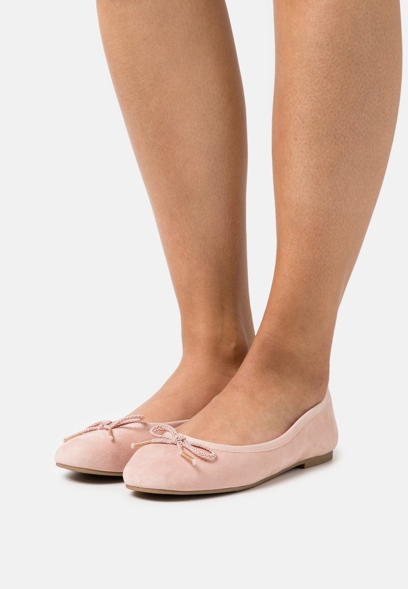 Jambes portant des ballerines en daim rose clair avec de petits nœuds sur le dessus, se tenant sur un fond clair uni.