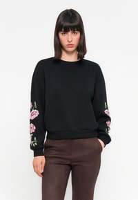 Mustame sweatshirt, millel on roosa ja rohelise lilleline tikand varrukatel, ribatud äär ja mansetid, lõdvem sobivus. Mudel kannab pruune pükse.